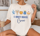 A woman wearing a cream-colored graphic t-shirt with a Hanukkah-themed design that reads '8 CRAZY NIGHTS Crew' and features cartoon illustrations of a driedel, menorah, latke, gelt and donut.