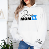 A woman wearing a white hoodie with a 'Dance MOM ZZ' graphic on the chest and 'Nikki' printed on the sleeve, paired with blue jeans, standing against a light wood paneled wall.