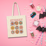 A cream canvas tote bag with 'SYDNEY' and a colorful skull-and-flower design, styled in a flat lay on a pink background with Halloween-themed props like paper bats, marshmallows, candy jars, and a candle.