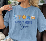 A woman wearing a blue graphic t-shirt with a Hanukkah-themed design that reads '8 CRAZY NIGHTS Crew' and features cartoon illustrations of a driedel, menorah, latke, gelt and donut.