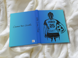 A custom hardcover photo book lying open on a textured white knit blanket. The cover features a blue gradient design with a black line-art illustration of a soccer player from behind holding a ball, with the name 'JONES' and number '25' printed on the back.