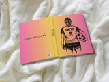 A hardcover journal or notebook shown open on a textured white knit blanket. The cover features a pink-to-yellow gradient with a black graphic of a soccer player from behind holding a ball, the name 'SMITH' and number '9' on the jersey, the year '2026' on the spine, and the phrase 'Chase the Goal.