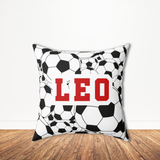 A square throw pillow with a black and white soccer ball pattern and the name 'LEO' printed in bold red letters, displayed on a wooden floor against a white wall.