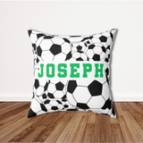 A square throw pillow with a black and white soccer ball pattern and the name 'JOSEPH' printed in green across the center, set on a wooden floor against a white wall.