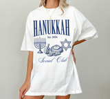 A woman wearing an oversized white graphic T-shirt with blue text reading 'HANUKKAH Est. 2026 Social Club' and illustrations of a menorah, dreidel, and Star of David, against a plain light gray background.