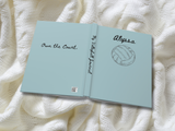 An open hardcover journal titled 'Own the Court.' lies on a textured cream knit blanket.