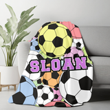 A colorful throw blanket draped over a gray sofa in a living room setting, featuring soccer ball patterns and the name 'SLOAN' in large purple letters. A potted plant is visible on the left.
