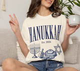 A woman sitting on a light beige couch wearing a cream-colored graphic t-shirt with 'HANUKKAH Est. 2026' printed in navy blue, along with illustrations of a menorah, dreidel,  and Star of David.