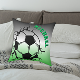 A square decorative throw pillow on a bed, featuring a graphic of a soccer ball bursting through cracks with the name 'MICHAEL' printed in green.