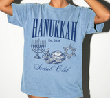 A person wearing a light blue short-sleeve graphic T-shirt with 'HANUKKAH Est. 2026 Social Club' text and Hanukkah-themed illustrations 