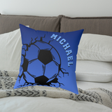 A square decorative throw pillow on a bed, featuring a graphic of a soccer ball bursting through cracks with the name 'MICHAEL' printed in blue.