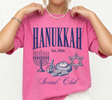 A person wearing a bright pink graphic T-shirt with navy text reading 'HANUKKAH Est. 2026 Social Club' and illustrations of Hanukkah symbols including a menorah, dreidel, matzah, gelt, and Star of David.