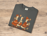 A dark gray folded t-shirt with a graphic of three cartoon geese wearing orange sweaters and hats while holding mahjong tiles, displayed on a light brown wooden background.