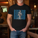 A man wearing a black graphic t-shirt with a blue and white 'Dance Dad' design, with a black and white photo of his daughter dancing, with her name in cursive, standing in a rustic indoor setting.