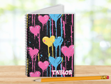 A spiral-bound notebook with a black cover decorated with colorful paint-drip hearts in pink, blue, and yellow, and the name 'TAYLOR' printed at the bottom. The notebook stands on a wooden surface with a yellow pencil in front, against a softly blurred outdoor background.