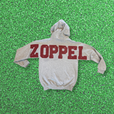 A light gray hoodie with 'ZOPPEL' printed across the back in distressed dark red block letters, laid flat on a green artificial turf background.