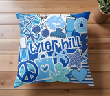 A square decorative throw pillow with a blues and white graphic collage featuring the camp 'tyler hill' in large blue letters, hearts, stars, and candy illustrations, resting on white bedding.
