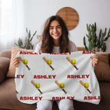 a girl holding a personalized white blanket with a repeating pattern of red 'ASHLEY' text, yellow softballs, and wooden bats.