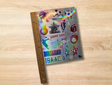  a sheet of colorful sports-themed stickers. The sheet features cartoon-style sports icons including a basketball, soccer ball, football, baseball, glove, star, and lightning bolts, with the text 'GAME DAY' and 'ISAAC' repeated. The sheet is up against a ruler to show it is 9 inches long. 