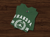 A folded green t-shirt with a white graphic print reading 'GRANDPA 2026' and a laurel wreath, displayed on a dark wood background.