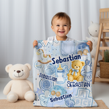 A smiling toddler stands behind a personalized baby blanket featuring blue and white illustrations and the name 'Sebastian' repeated, with a cream teddy bear beside them in a softly lit nursery setting.