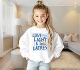 A young girl with a high ponytail stands in a bright, modern living room wearing a white crewneck sweatshirt with a blue and yellow graphic that reads 'LOVE LIGHT & LATKES' 