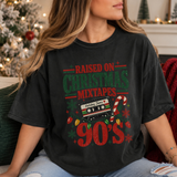 A woman wearing a black graphic t-shirt with a retro 90s Christmas cassette tape design that reads 'RAISED ON CHRISTMAS MIXTAPES 90's'. The photo is taken in a cozy living room setting with a decorated Christmas tree in the background.