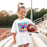 Little girl standing in bleachers holding a football. She's wearing a white t-shirt with a colorful design: teal vertical stripes, pink, purple, yellow and orange footballs scattered around. Inside the balls are lighting bolts, flowers, hearts, leopard print, etc. At the bottom in pink and purple it says Football Sister.