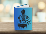A hardcover notebook with a bright blue cover standing on a wooden surface. The back of the cover features a black graphic illustration of a soccer player from behind, wearing a jersey with the name 'JONES' and number '25', while holding a soccer ball.