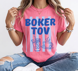A woman wearing a pink graphic t-shirt with blue text reading 'BOKER TOV' and a drink and coffee cup illustration, paired with distressed blue jeans and white sneakers.