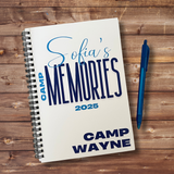 A spiral-bound notebook titled "Sofia's Memories 2025" with "Camp Wayne" printed on the cover, placed on a rustic wooden surface next to a blue pen.