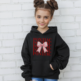 A young girl with her hair in two buns wears a black hoodie featuring a red repeating 'PONTIAC' graphic and a pink bow design on the front. She is standing against a white brick wall.