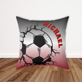 A square decorative throw pillow featuring a soccer ball graphic with a crackle effect, set against a gray-to-red gradient background with the name 'MICHAEL' in red text, shown standing on a wooden floor against a white wall.