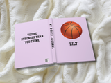 A personalized basketball-themed journal shown open on a textured white knit blanket. The cover is light lavender pink, featuring a graphic of an orange basketball and the name 'LILY' printed in black.