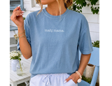 A woman wearing a light blue short-sleeve t-shirt with the text 'mahj mama.' printed in white across the chest, paired with white and blue striped drawstring shorts, set against a bright outdoor patio background.