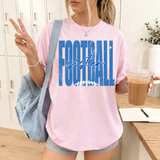 A woman wearing a light pink graphic t-shirt with 'FOOTBALL sister' printed in blue across the front, holding an iced coffee and posing in front of a locker room background.
