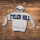 A light heather gray pullover hoodie with 'TYLER HILL' printed across the chest in dark  blue varsity block letters, laid flat on a rustic brown wooden plank background.