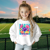 Little girl standing in bleachers holding a football. She's wearing a white crewneck sweatshirt with a colorful design: teal vertical stripes, pink, purple, yellow and orange footballs scattered around. Inside the balls are lighting bolts, flowers, hearts, leopard print, etc. At the bottom in pink and purple it says Football Sister.