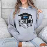 A woman wearing a heather gray hoodie with a custom black and blue graphic that reads 'LAYLA SENIOR 2026' with a graduation cap, volleyball, and heart design, sitting in a cozy room setting.