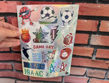 A hand holds a sheet of colorful sports-themed stickers against a red brick wall background. The sheet features cartoon-style sports icons including a basketball, soccer ball, football, baseball, glove, star, and lightning bolts, with the text 'GAME DAY' and 'ISAAC' repeated.