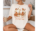 A woman sitting on a bed wearing an oversized cream-colored t-shirt with a graphic of three geese in orange sweaters and hats holding mahjong tiles, with the text 'IT'S TILE TIME' printed below. She is holding an iced coffee in a cozy bedroom setting.
