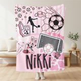 A pink fleece throw blanket with a personalized soccer theme, featuring the name 'Nikki' in black letters, soccer balls, a net, a foaming hand, and a player silhouette, held up in a living room setting.