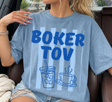 A woman wearing a light blue graphic t-shirt with the text 'BOKER TOV' and illustrations of an iced coffee cup and a hot coffee cup on a striped background, sitting in a car.
