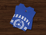 A folded royal blue t-shirt with a white collegiate-style 'Grandpa 2026 Champion' laurel wreath graphic, displayed on a dark wood background.