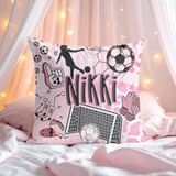 A pink throw pillow with a soccer-themed graphic print featuring the name 'Nikki' in large black letters, surrounded by soccer illustrations like balls, a goal net, gloves, and a fan sign. The pillow is styled on a bed under pink drapes with warm string lights.