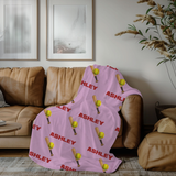 A personalized pink blanket with a repeating pattern of red 'ASHLEY' text, yellow softballs, and wooden bats, laid over a leather couch. 