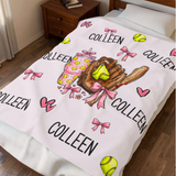 A personalized softball-themed blanket featuring the name 'COLLEEN' repeated, decorated with illustrations of pink bows, pink hearts, yellow softballs, a brown softball glove, and a pink drink cup, displayed on a light wood plank background.
