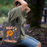 A woman sitting outdoors on a bench carries a black tote bag with a colorful pumpkin graphic and the text 'RILEY'S TREATS'.