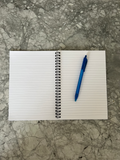 A white lined spiral notebook lies open on a gray marble surface, with a blue pen placed on the right side of the page.