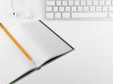 An open lined notebook with a yellow pencil on top, styled on a white desk alongside a white keyboard and earbuds.